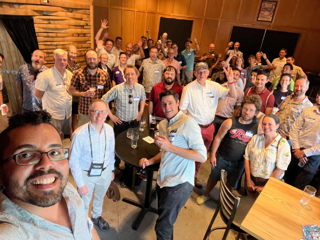 The image is a high-angle group selfie featuring a large group of people gathered inside what appears to be a casual, social setting, possibly a bar or pub. Most of the individuals are standing and facing the camera with a variety of happy expressions; many are smiling, waving, or holding up their drinks in a toasting gesture. Everyone seems to be enjoying the occasion. The person taking the selfie is at the bottom left of the image, holding the camera with one hand and smiling broadly. The attendees are dressed in a mix of casual and business casual attire, suggesting a relaxed but possibly professional gathering. The environment has a rustic vibe with a wood-paneled wall in the background, enhancing the informal atmosphere of the event.