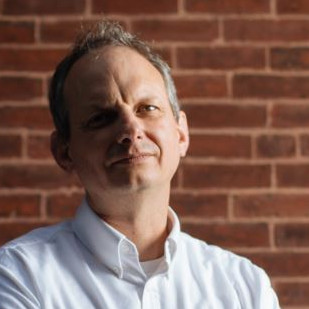 Headshot photo of Evan Manvel, who is posed in front of a brick wall.