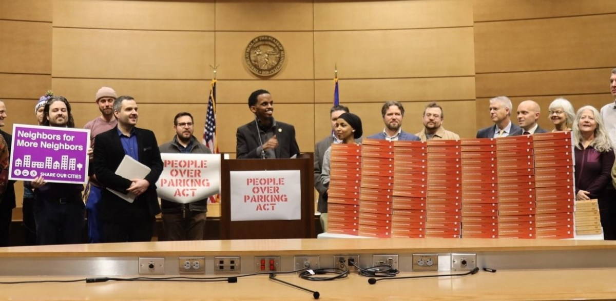 This image shows a group of people gathered in what appears to be a government building or a public meeting hall. In the foreground, a man stands at a podium with a sign reading "PEOPLE OVER PARKING ACT". He is flanked by others holding signs, such as "Neighbors for More Neighbors" with additional text that is not fully legible. To the right, there is a tall stack of red-colored binders or documents, labeled as the "Impact Report 2023". This suggests that the event is related to urban planning or housing policy, possibly advocating for prioritizing community and living space over car parking areas. There are U.S. flags in the background, indicating this may be in the United States. The atmosphere suggests a formal announcement or advocacy effort with media presence, as the event seems organized for a significant announcement or report release.