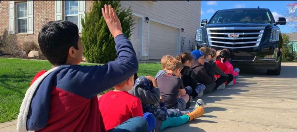 Picture of a modern Cadillac Escalade with 13 kids sitting in front of it.