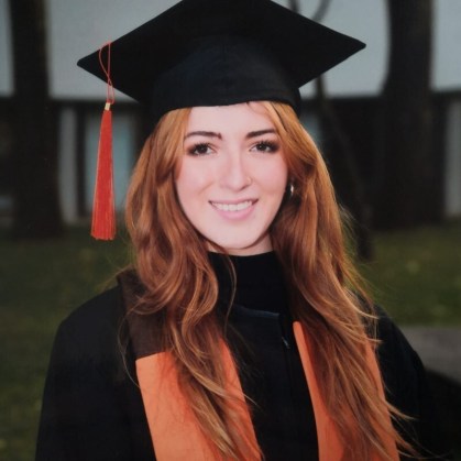 Headshot photo of PRN's new full-time member, Paula Acero. She is smiling in her graduation garb. In the background is grass and a building.
